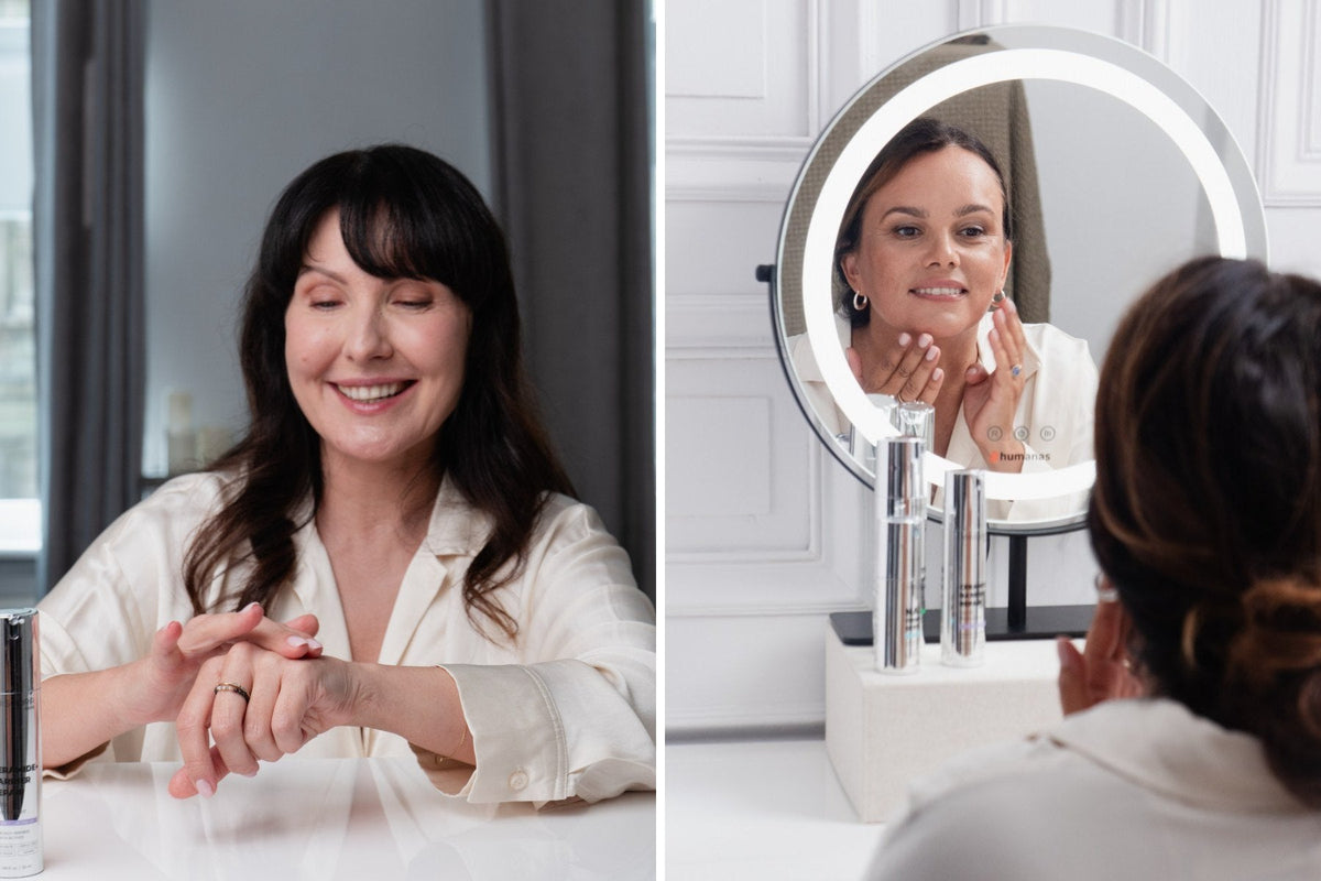 Woman in her 50s smoothing cream over the back of her hand beside a woman checking her skin in a lit vanity mirror