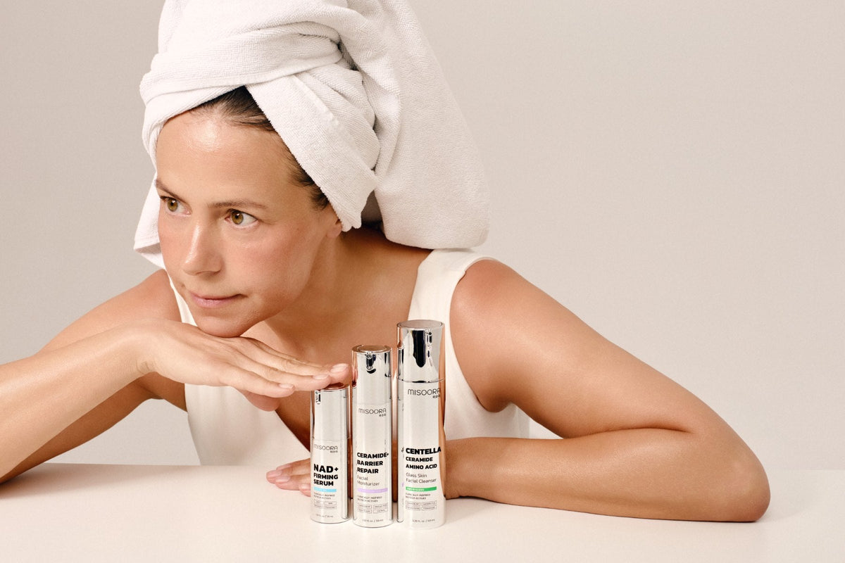 Woman in her 50s with a towel on her head after a shower, resting her chin on her hand beside three MISOORA bottles