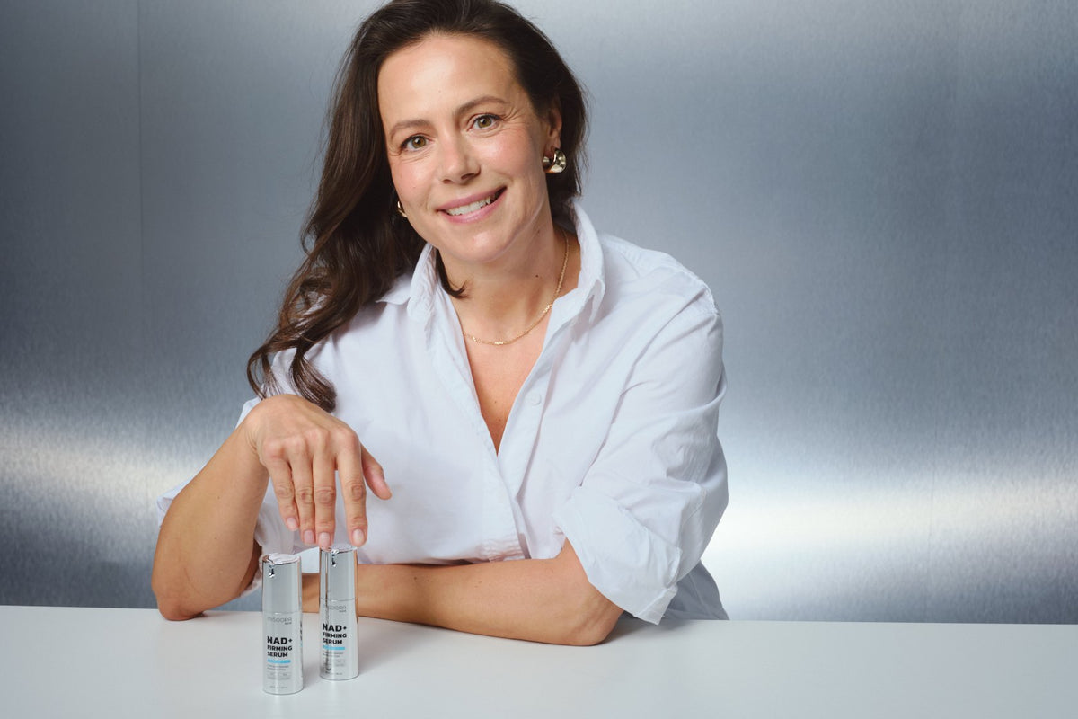 Smiling woman in her 50s in a white shirt at a table with MISOORA NAD+ serum